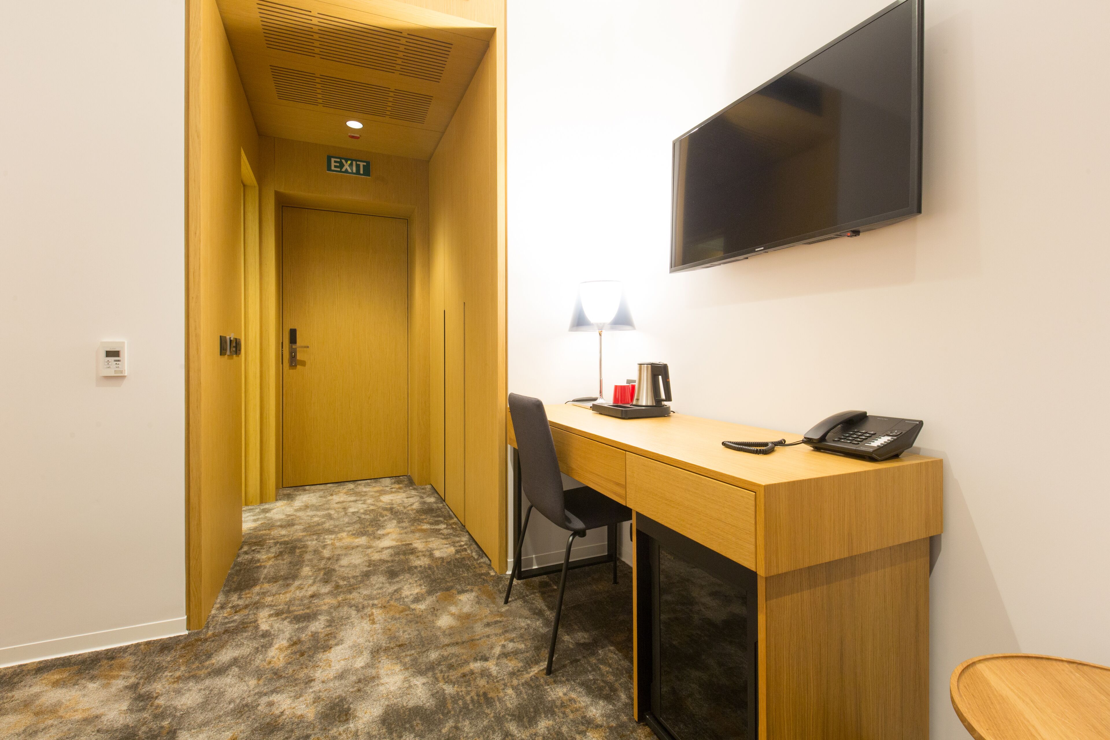 A hotel room with a desk, chair, lamp, phone, and a TV mounted on the wall.