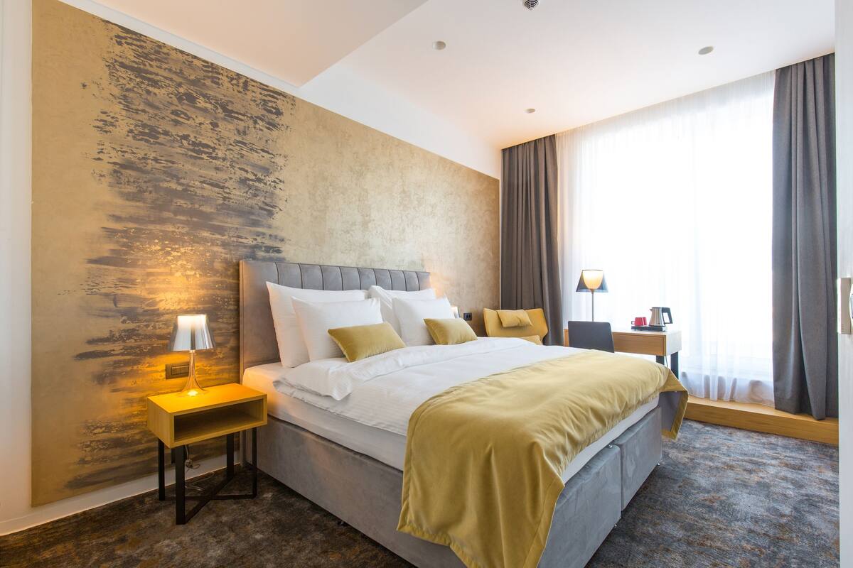 A modern hotel room with a large bed, a nightstand with a lamp, a wooden side table, and a view of the outdoors through a window with curtains.
