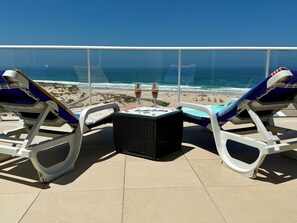 A balcony with two sun loungers, a small table, and a glass of wine. The view includes a beach and the ocean. - Luxury Penthouse with 180° Ocean and 270° Panoramic Views, Praia d'El Rey, Silvercoast (Vale de Janelas)