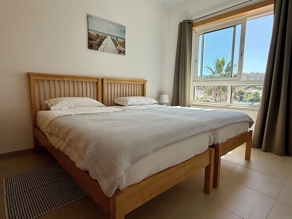 A bedroom with a wooden bed, a view of the outdoors through a window, and a painting on the wall. - Luxury Penthouse with 180° Ocean and 270° Panoramic Views, Praia d'El Rey, Silvercoast (Vale de Janelas)