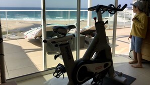 Ein Zimmer mit großem Fenster mit Blick auf einen Strand, einem stehenden Ergometer und einem Kind, das am Fenster steht.