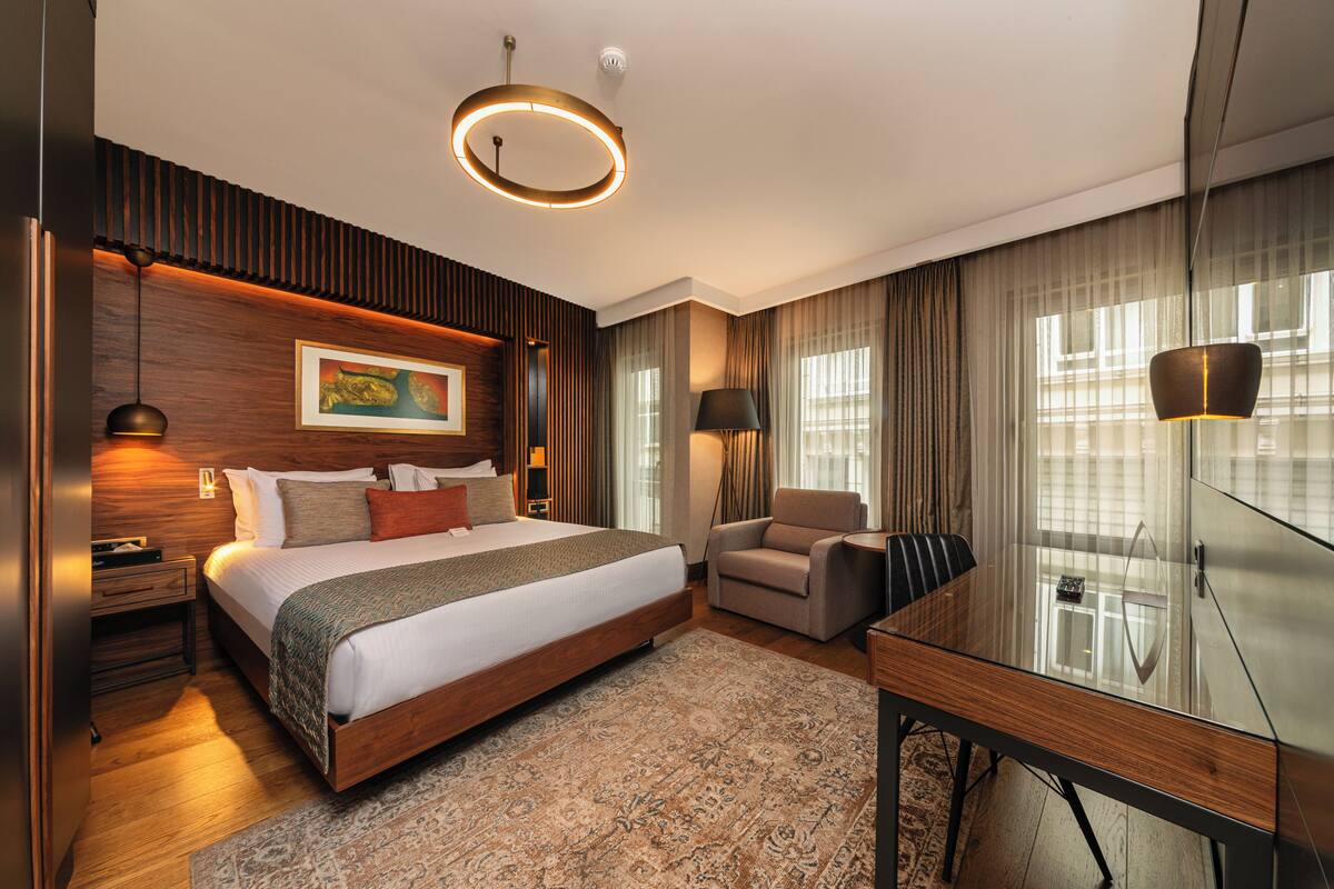 A modern hotel room with a large bed, a desk, a chair, and a glass cabinet.