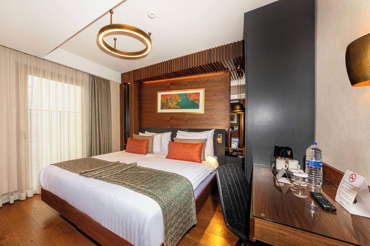 A hotel room with a large bed, wooden headboard, a desk with a chair, a desk lamp, a water bottle, a remote control, and a painting on the wall.