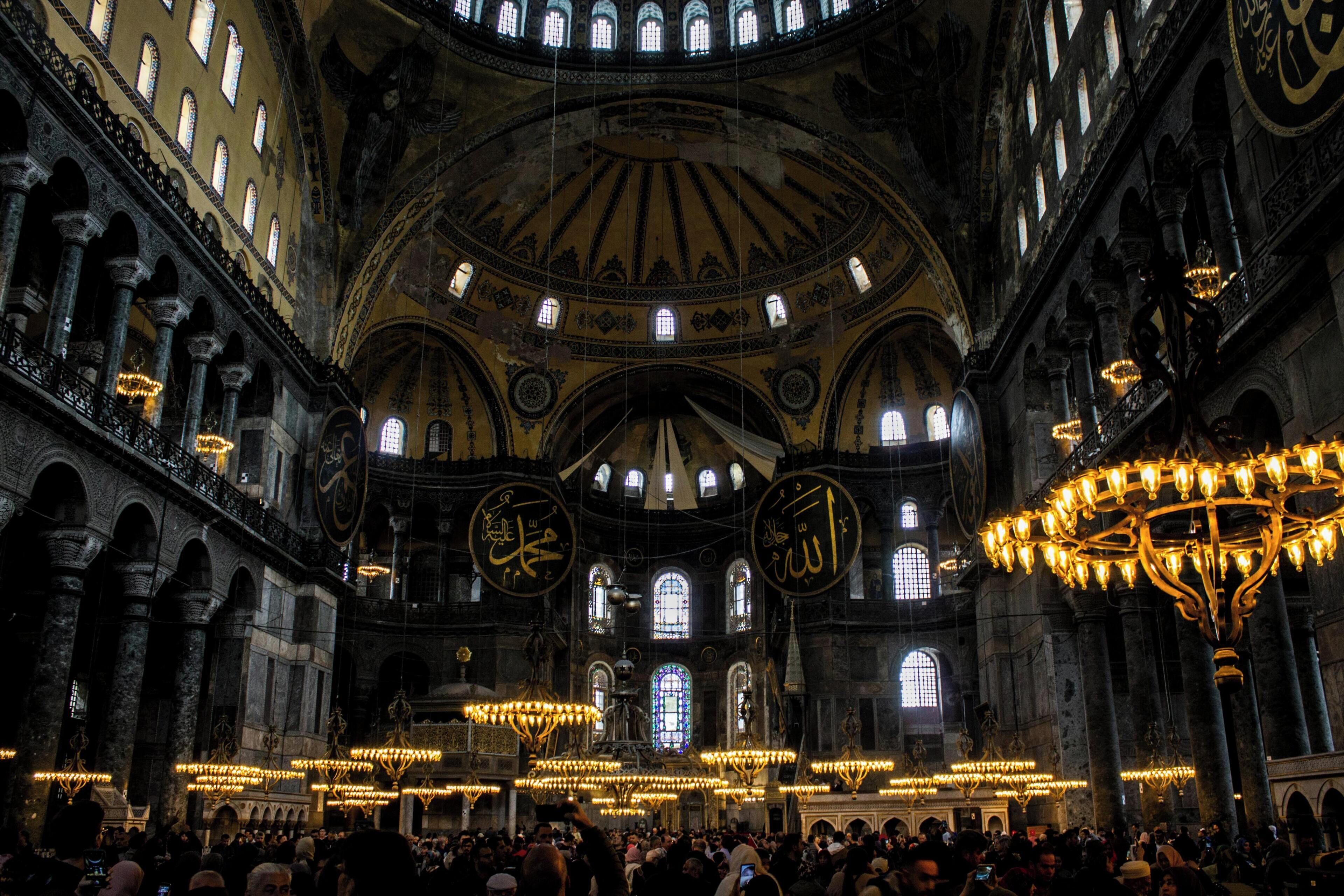 A crowded interior with high ceilings, arched windows, and ornate chandeliers.
