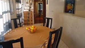 Un comedor con una mesa redonda, sillas y un frutero. Se ve una escalera con barandilla y una puerta que conduce a otra habitación.