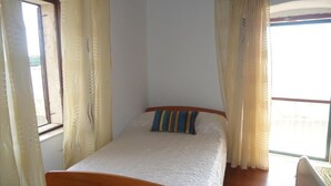 Una cama individual con cabecera de madera en una habitación con dos ventanas, cada una con cortinas.