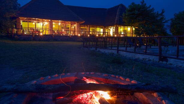 A lodge with a thatched roof, a well-lit dining area, and a fire pit in the foreground.