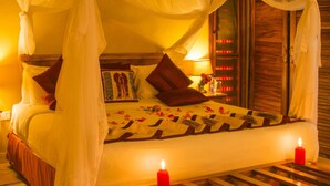 A bed with a canopy, pillows, a bedside table with a bottle and flowers, and a lit candle.