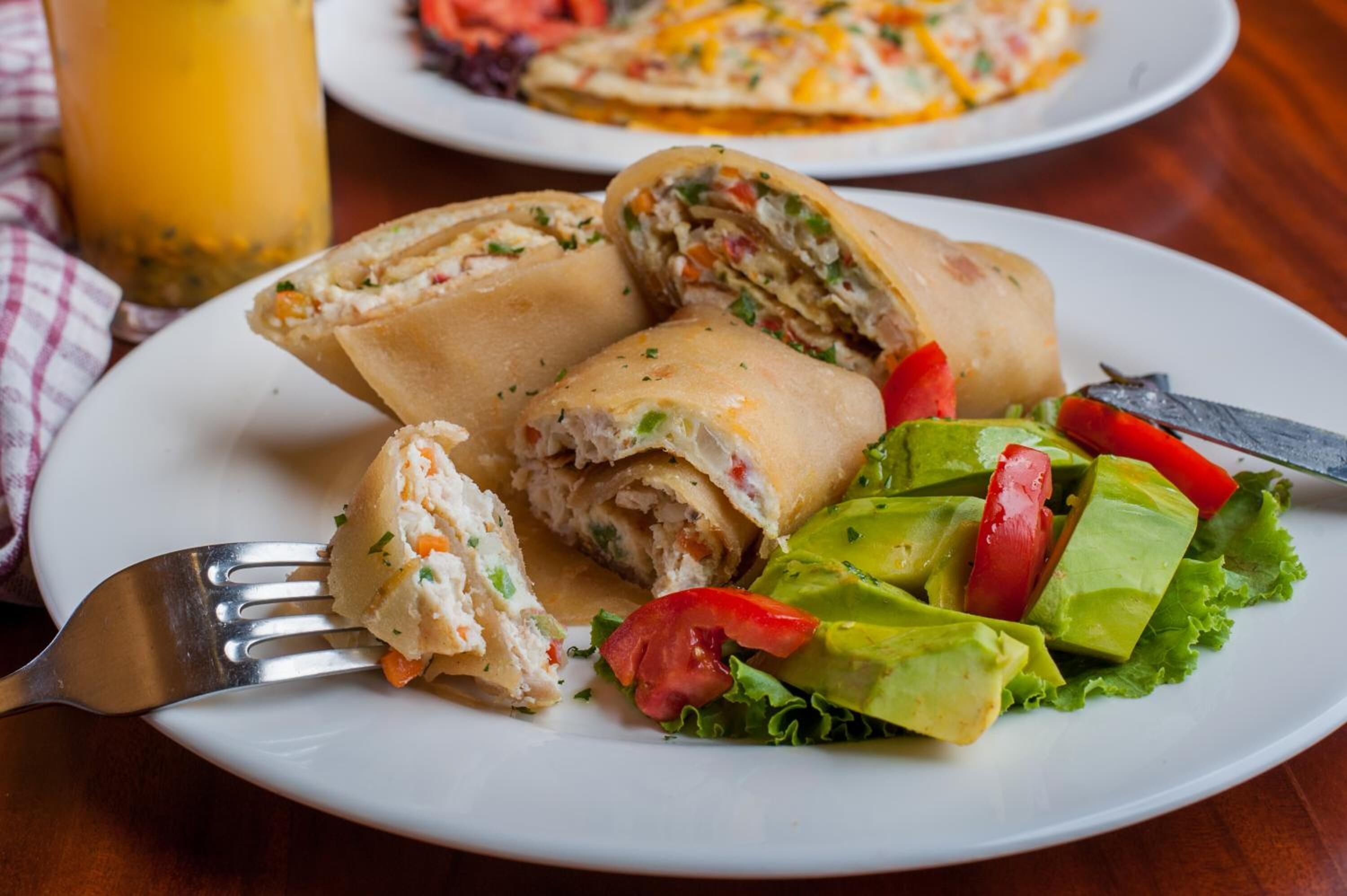 A plate with a rolled sandwich cut in half, a salad with avocado and tomatoes, and a glass of yellow beverage on a wooden table.