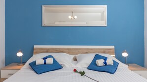 Una cama bien hecha con sábanas blancas y toallas azules, una rosa roja y dos lámparas de noche sobre mesitas de madera.