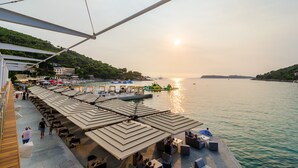 Un restaurante frente al mar con terraza y sombrillas, con vistas a un mar tranquilo y un paisaje montañoso.