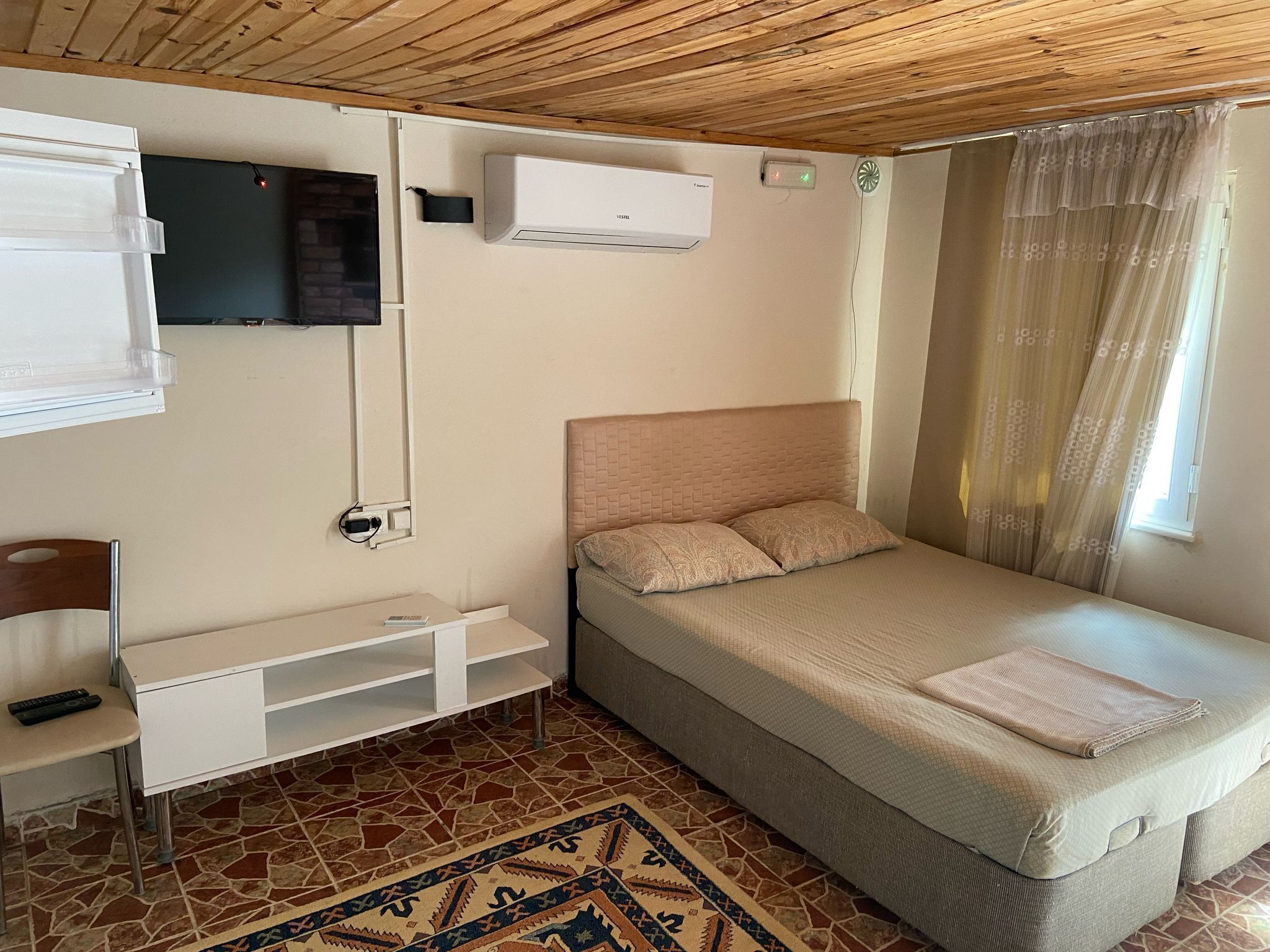 A single bedroom with a bed, a TV mounted on the wall, an air conditioning unit, and a small desk with a chair.