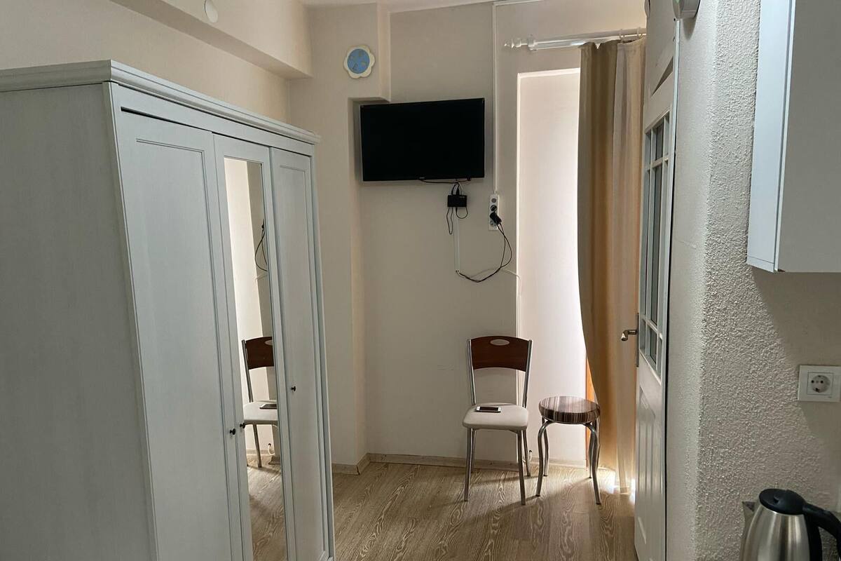 A room with a wooden floor, a white wardrobe, a television mounted on the wall, a chair, and a kettle on a table.