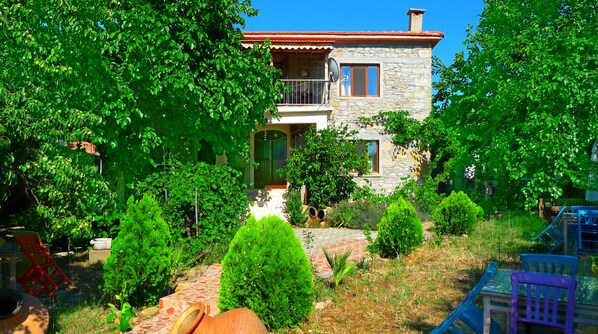 A stone house with a balcony, surrounded by lush greenery and a stone pathway. - Yesildag Pansiyon Gokceada (Gökçeada)