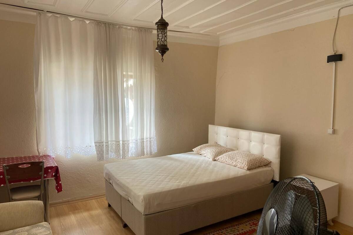 A bedroom with a bed, a desk, a chair, a standing fan, and a window with sheer curtains.
