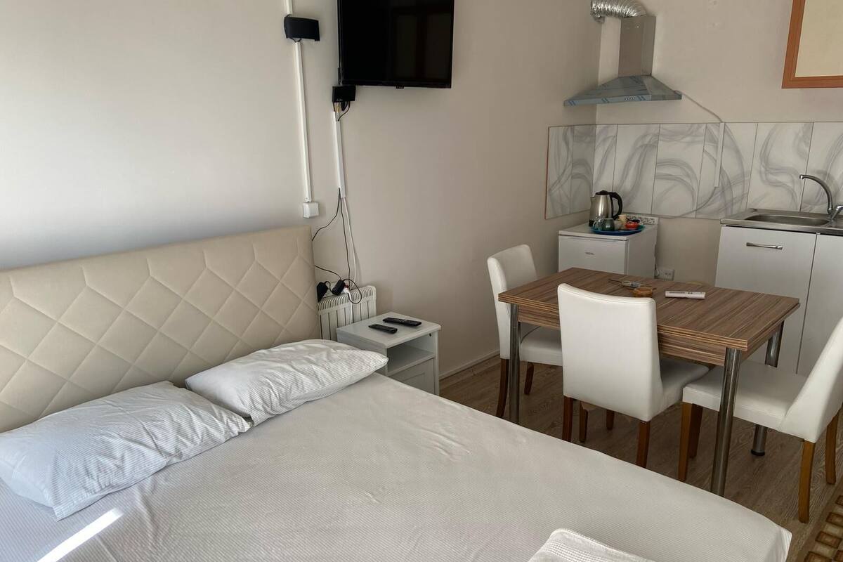 A hotel room with a bed, a desk, a chair, a small kitchenette, and a TV mounted on the wall.