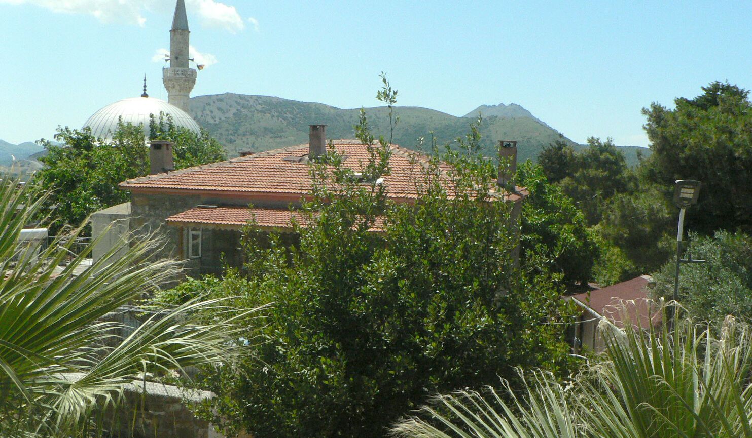Um edifício com telhado de telhas vermelhas, rodeado por vegetação e uma montanha ao fundo.