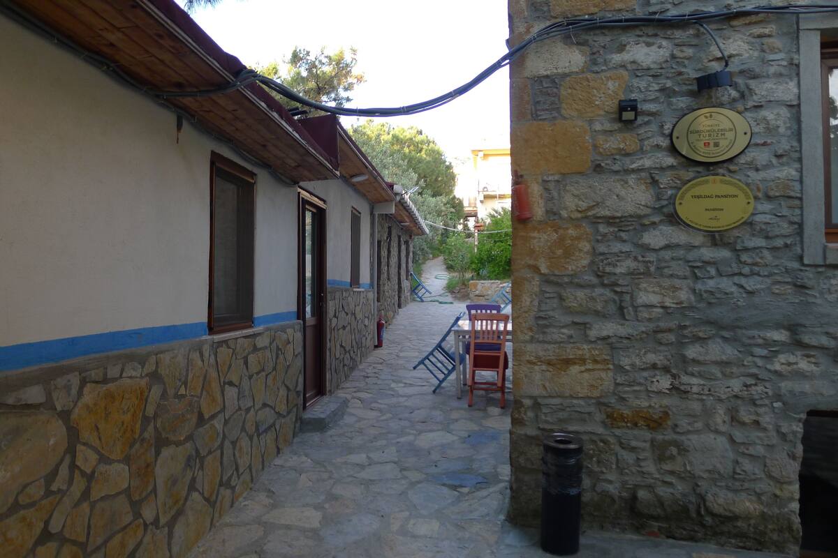 A narrow stone pathway leading to a building with a wooden roof and stone walls. There are two plaques on a stone wall, a fire extinguisher, and a chair outside.