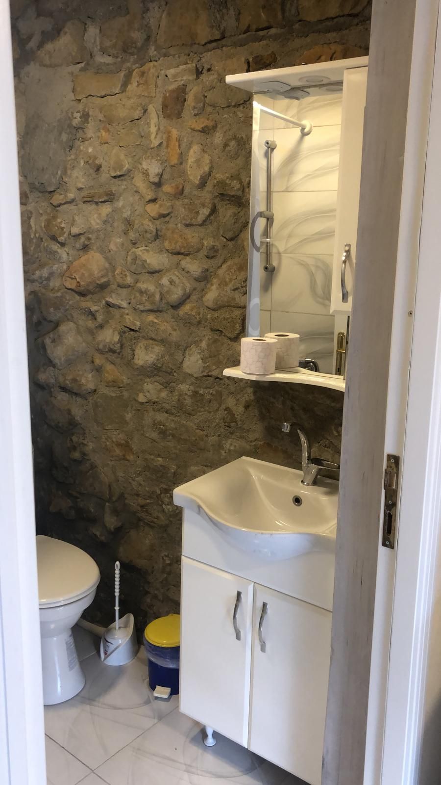 Un baño con pared de piedra, un lavamanos blanco, un inodoro y un armario para espejo.