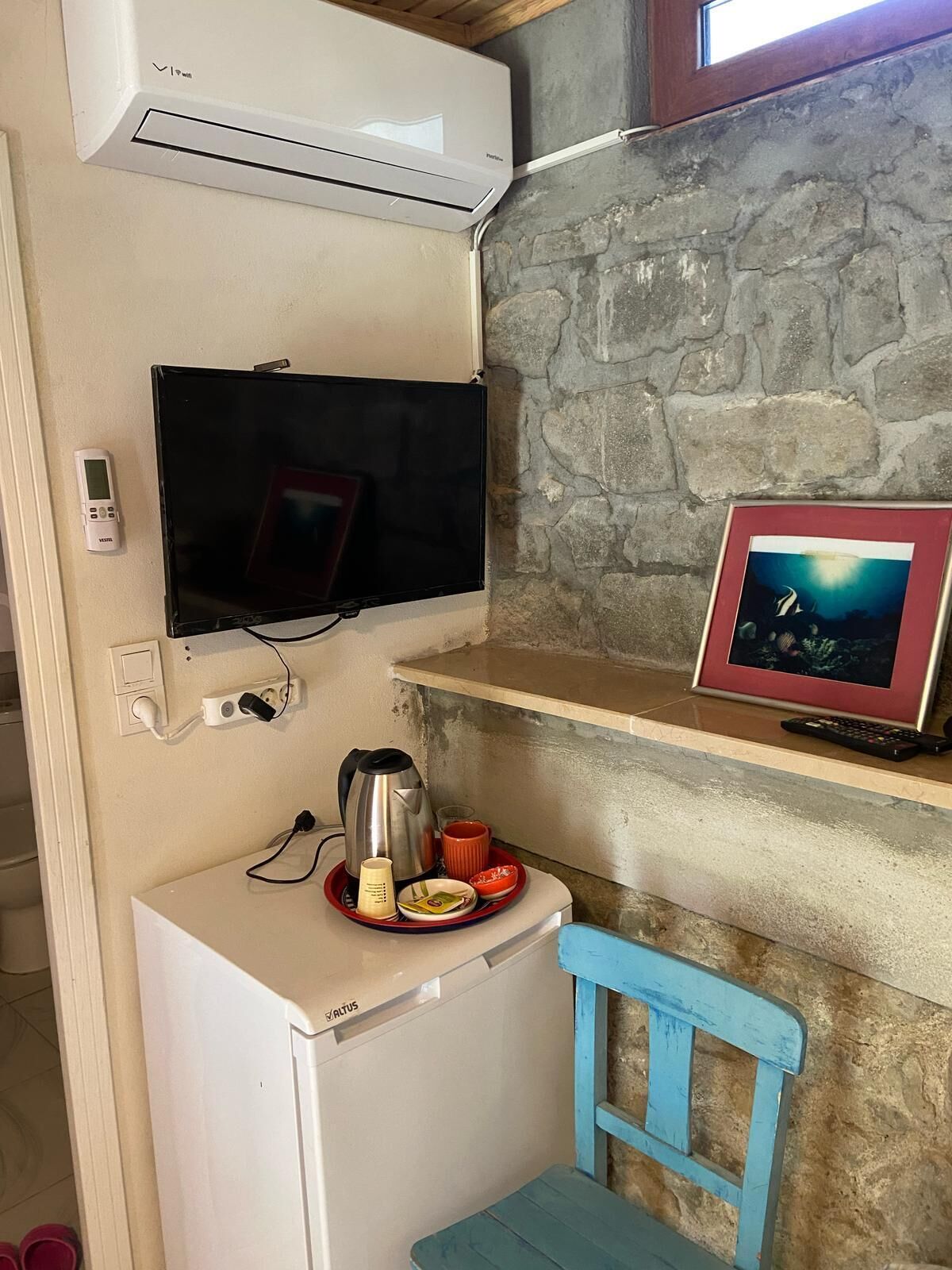 Una kitchenette compacta con un refrigerador blanco, una estufa pequeña, un televisor montado en la pared y un cuadro en un estante.