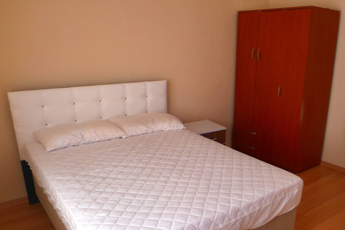 A neatly made bed with a quilted white duvet, a wooden nightstand, and a red wardrobe in a room with wooden flooring.