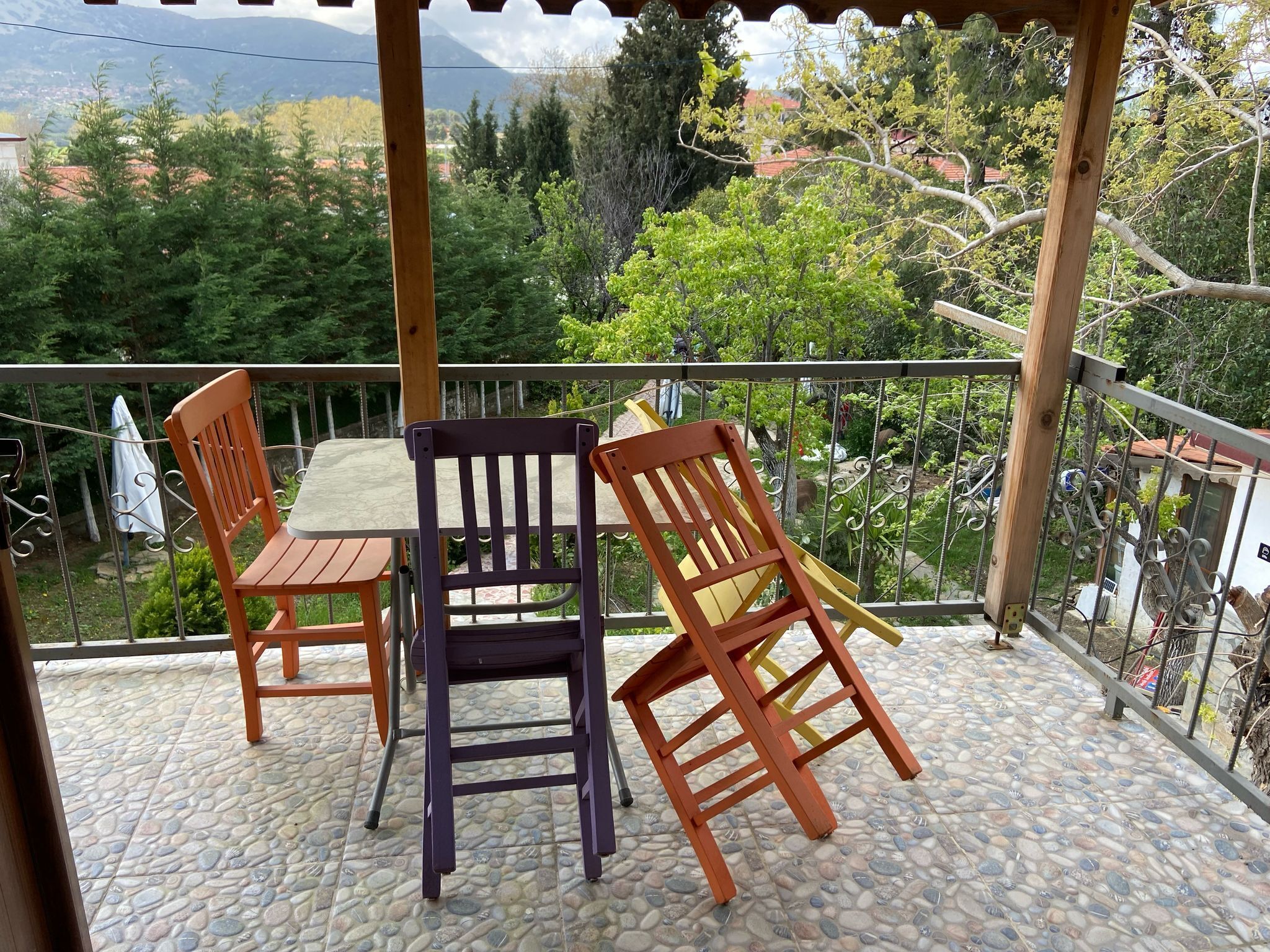 Un área de estar al aire libre techada con mesa y sillas, piso de baldosas de piedra y vista a un paisaje verde con árboles y montañas al fondo.