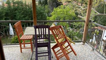 Un área de estar al aire libre techada con mesa y sillas, piso de baldosas de piedra y vista a un paisaje verde con árboles y montañas al fondo.