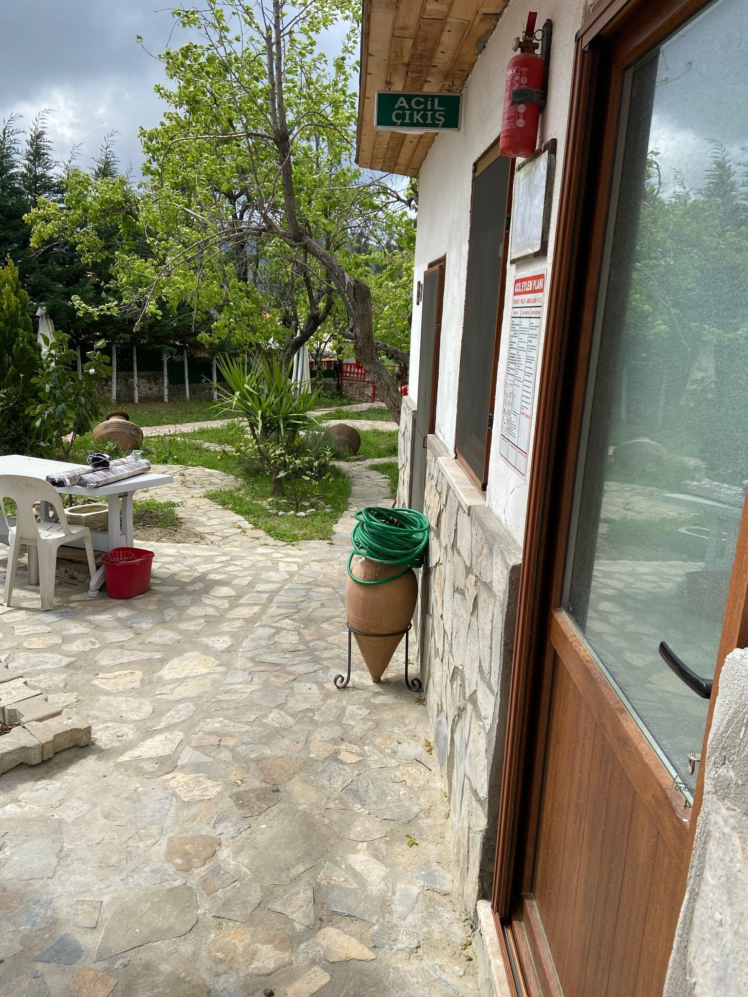 A stone-paved courtyard with a table and chairs, a large terracotta pot, and a fire extinguisher mounted on the wall.