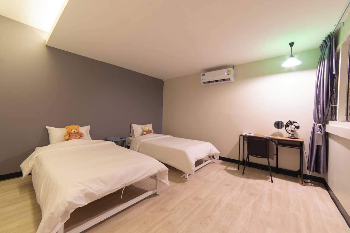Quarto de hotel com duas camas, uma escrivaninha, uma cadeira, aparelho de ar condicionado e janela com cortinas.