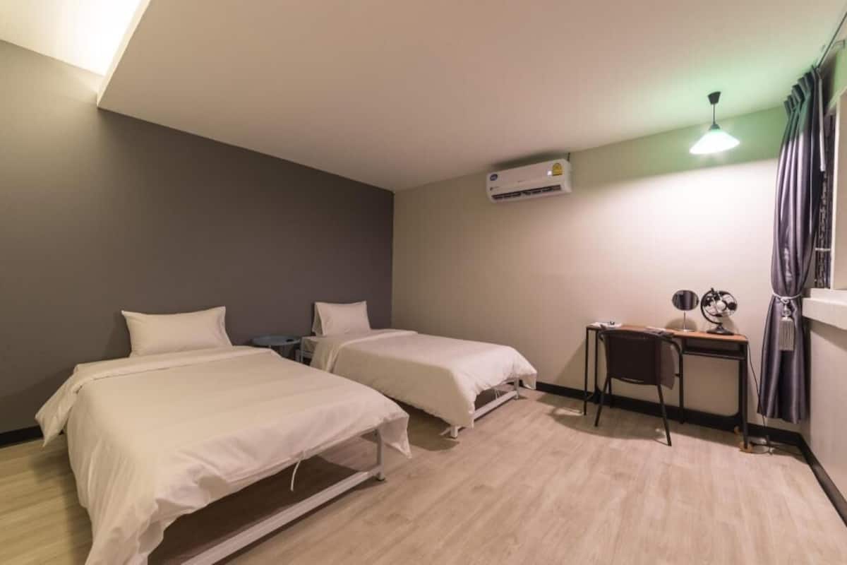 Quarto de hotel com duas camas, uma escrivaninha, uma cadeira, ar condicionado e uma janela com cortinas.