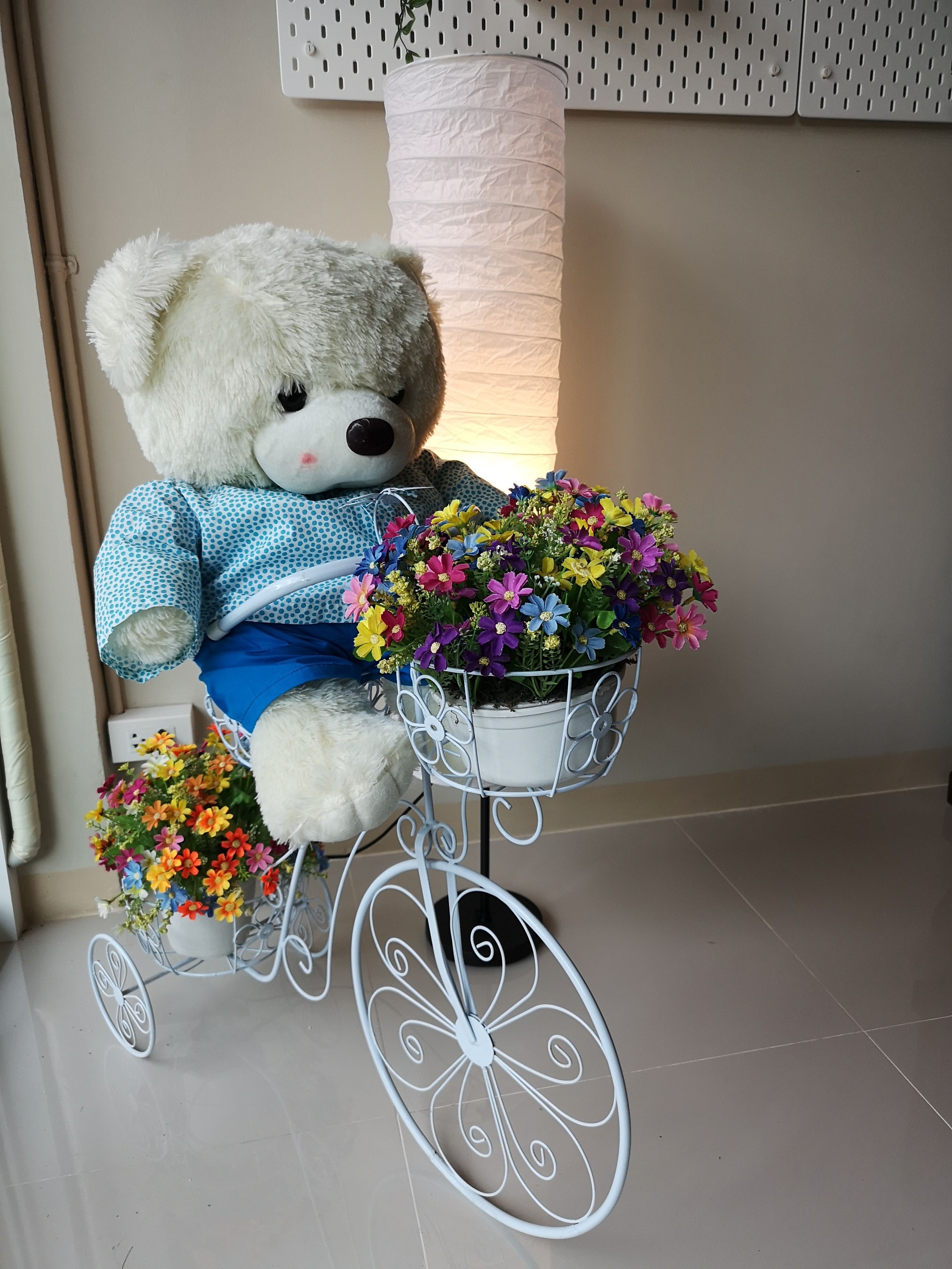 Un oso de peluche blanco sentado sobre una bicicleta metálica blanca con una cesta de flores.