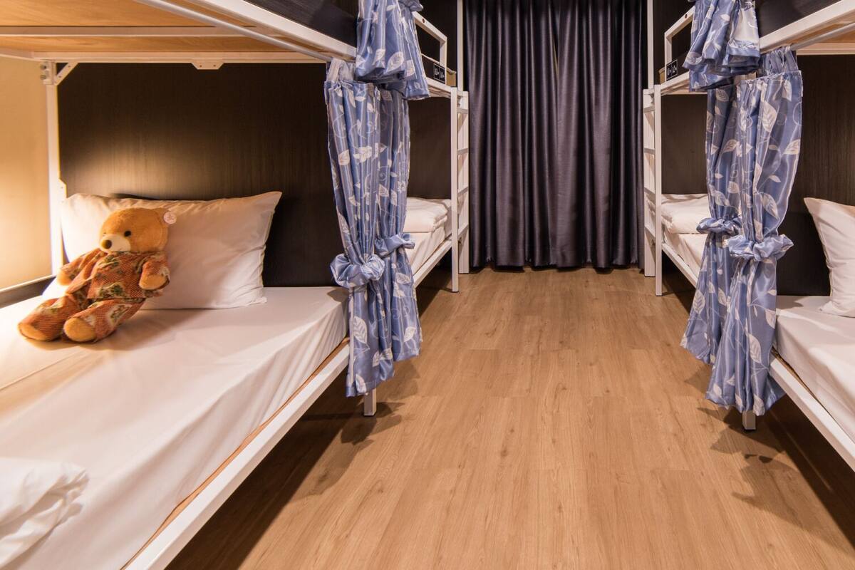 Quarto de hotel com duas camas de solteiro, cada uma com um ursinho de pelúcia, cortina e piso de madeira.