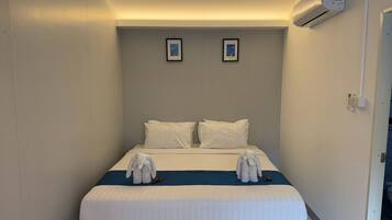 A hotel room with a bed, two pillows, a blue blanket, and a pair of white slippers on the bed. There is a wall-mounted air conditioner on the right side of the room.