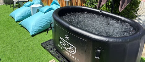 An outdoor ice bath with a wooden deck, blue cushions, and a wooden fence in the background.