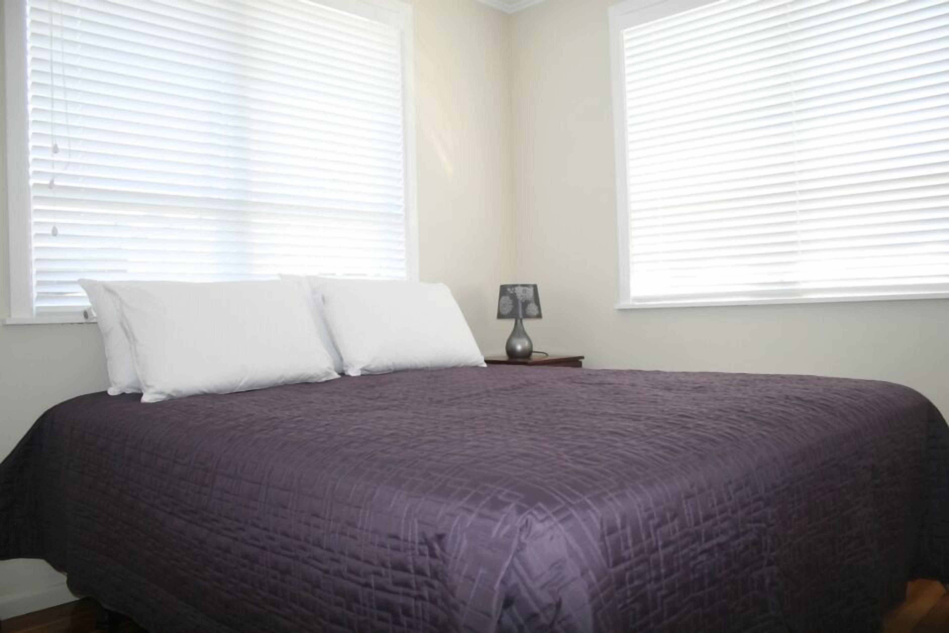 A bedroom with a large bed, two white pillows, a nightstand with a lamp, and two windows with blinds.