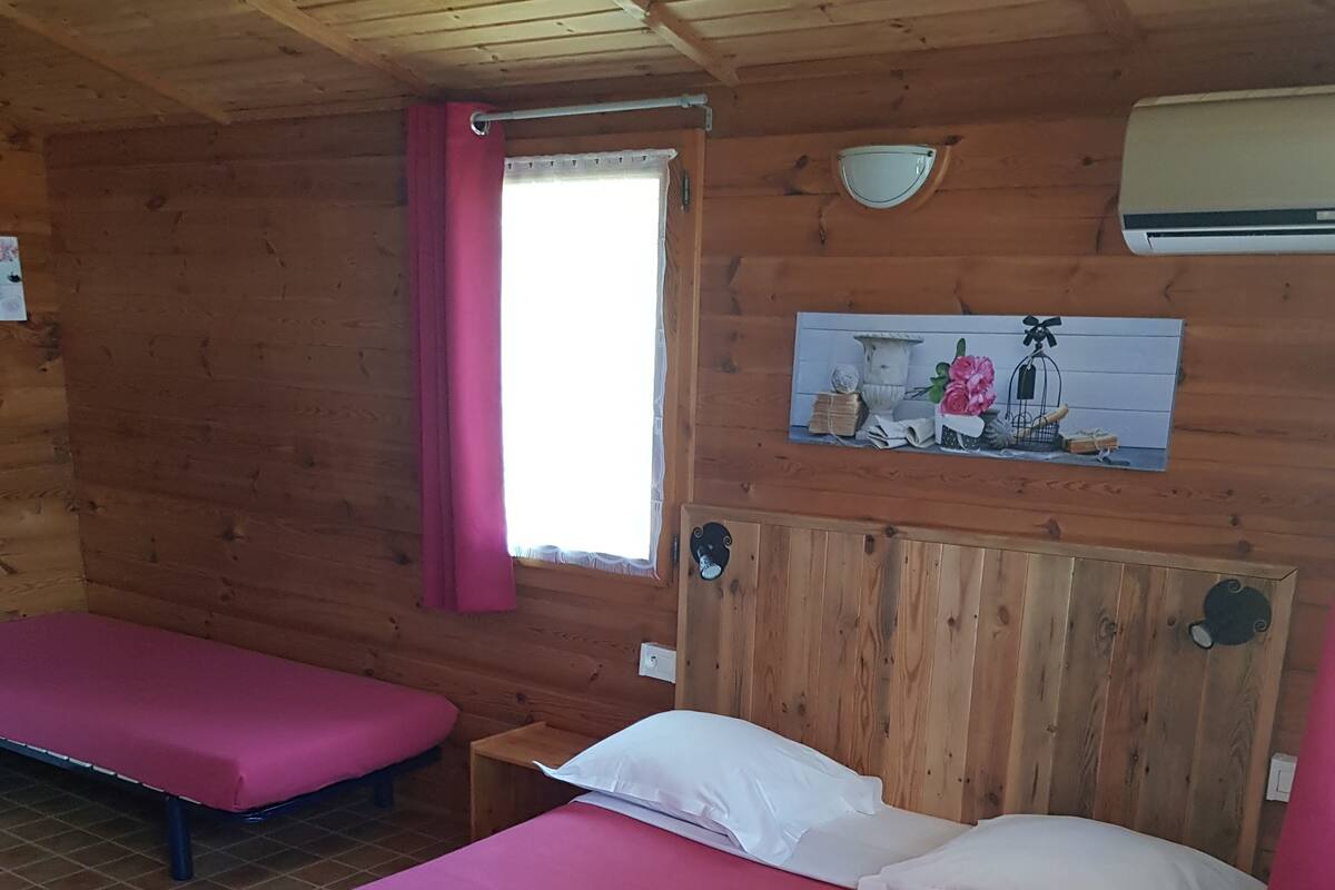Une chambre de chalet en bois avec deux lits, chacun recouvert d’une couverture rose et agrémenté d’oreillers blancs, et une fenêtre ornée d’un rideau rose.