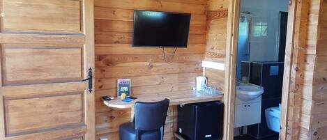 Habitación de cabaña de madera con escritorio, sillas, televisor y baño visible a través de una puerta abierta.