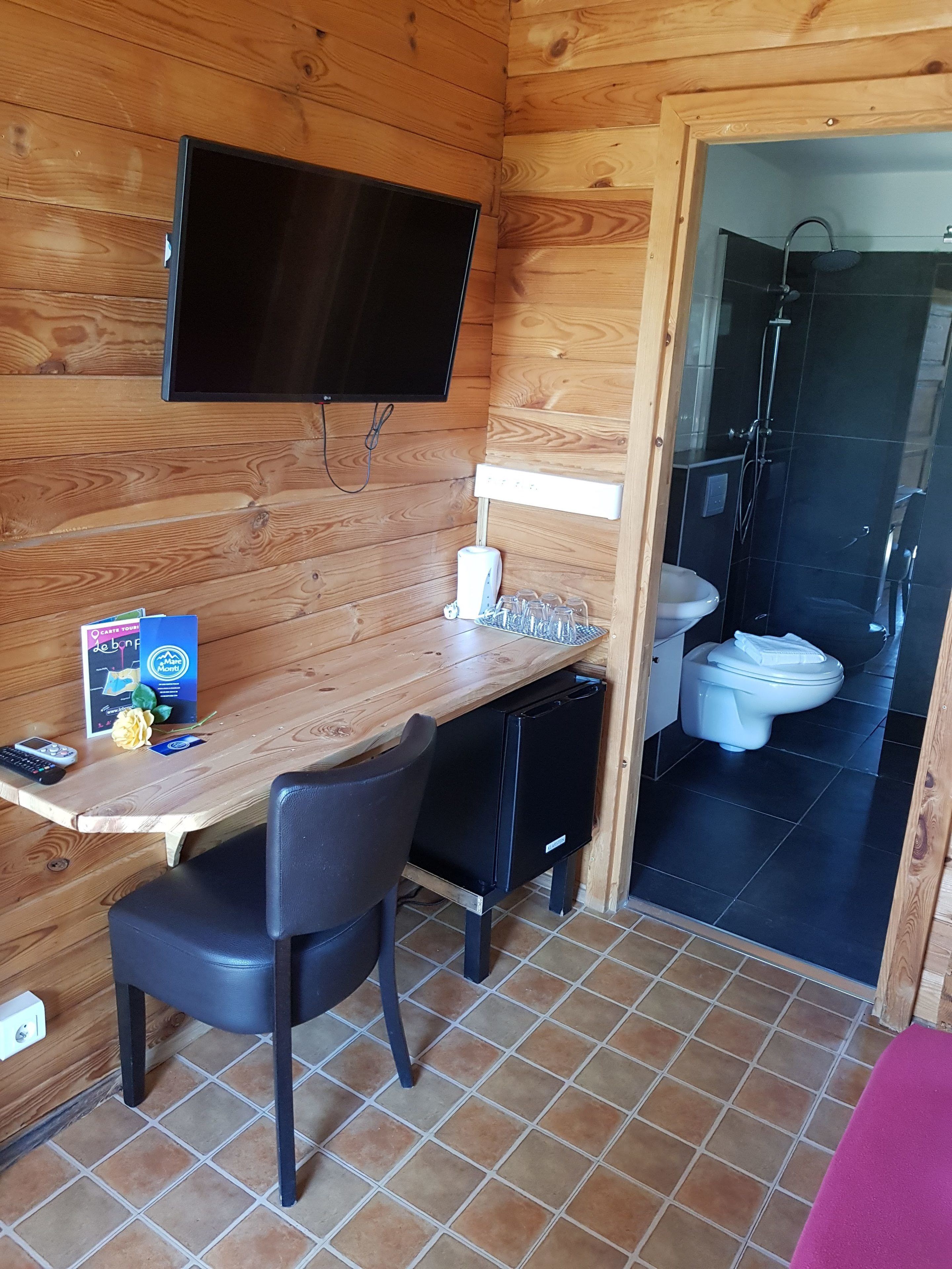 Een houten blokhut van binnen, met een houten bureau, een stoel, een aan de muur bevestigde televisie en een badkamer met douche en toilet.