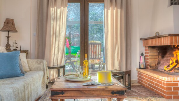 Ein gemütliches Wohnzimmer mit einer Couch, einem Tisch mit Kerze, einem Kamin und Blick durch eine Glastür.