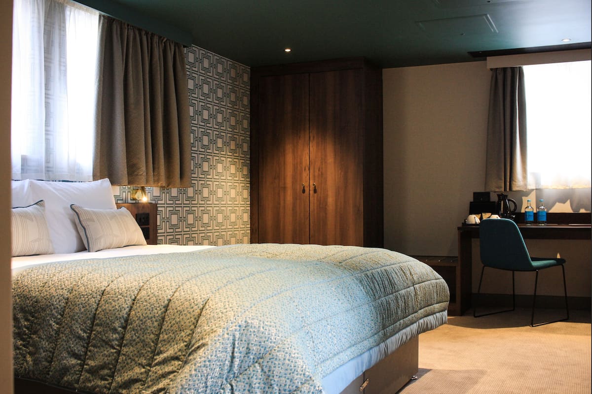 A hotel room with a large bed, a desk, a chair, and a wardrobe.