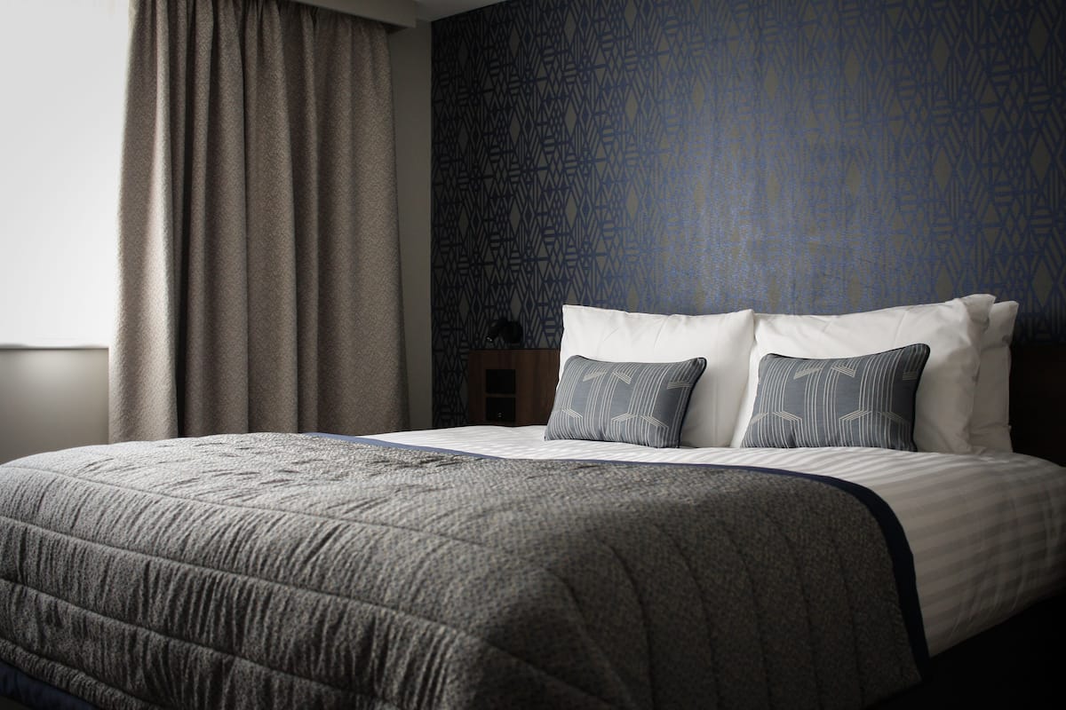 A neatly made bed with a grey quilt, two pillows with a geometric pattern, and a dark patterned wall behind.