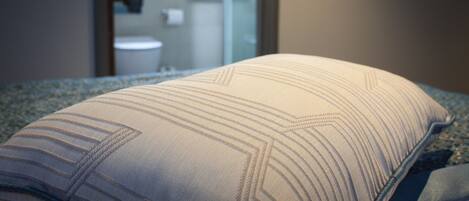 A close-up of a bed with a striped pillow, a bathroom in the background, and a glass shower door.