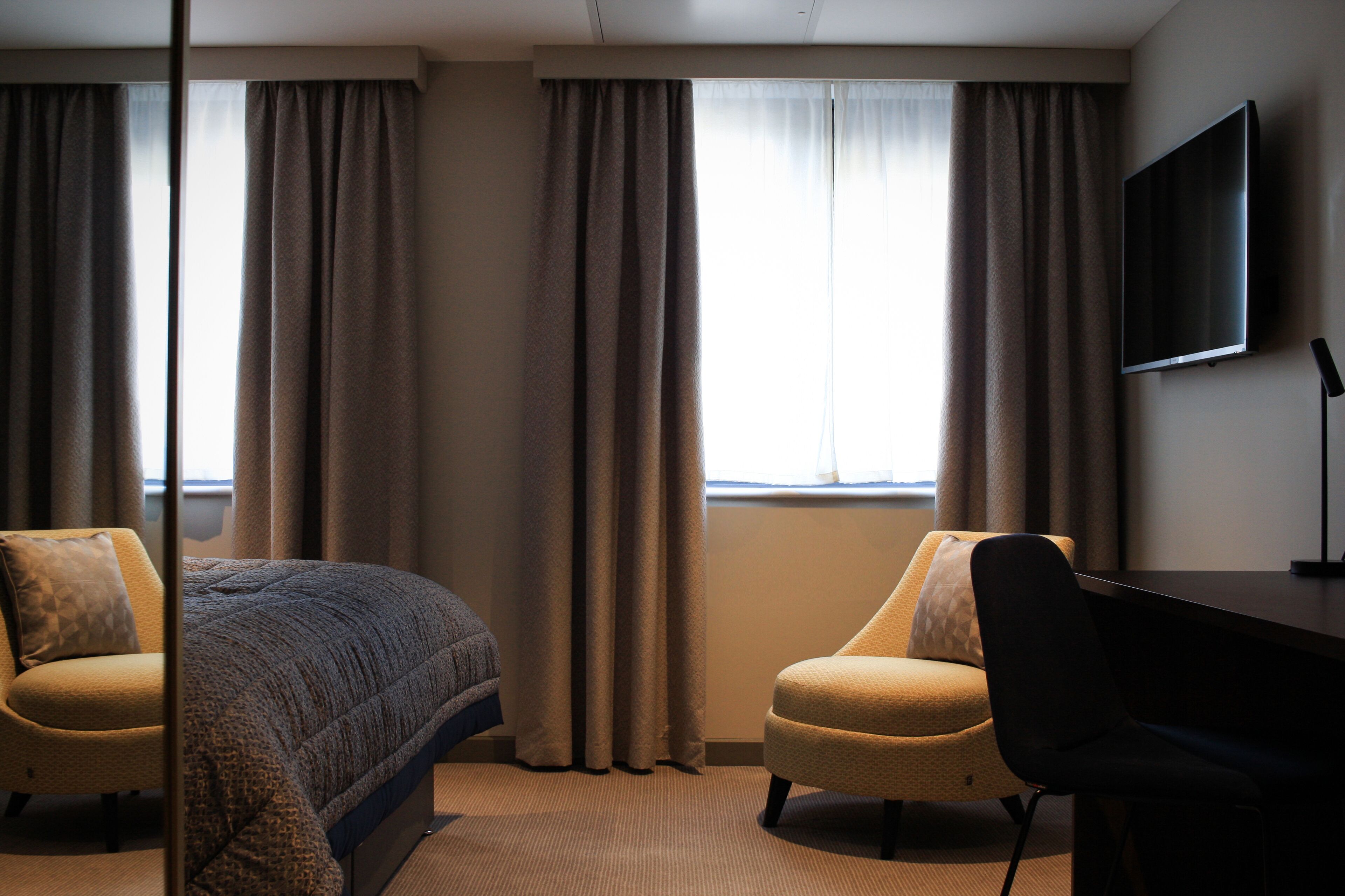 A hotel room with a bed, two armchairs, a desk, a TV, and large windows with curtains.