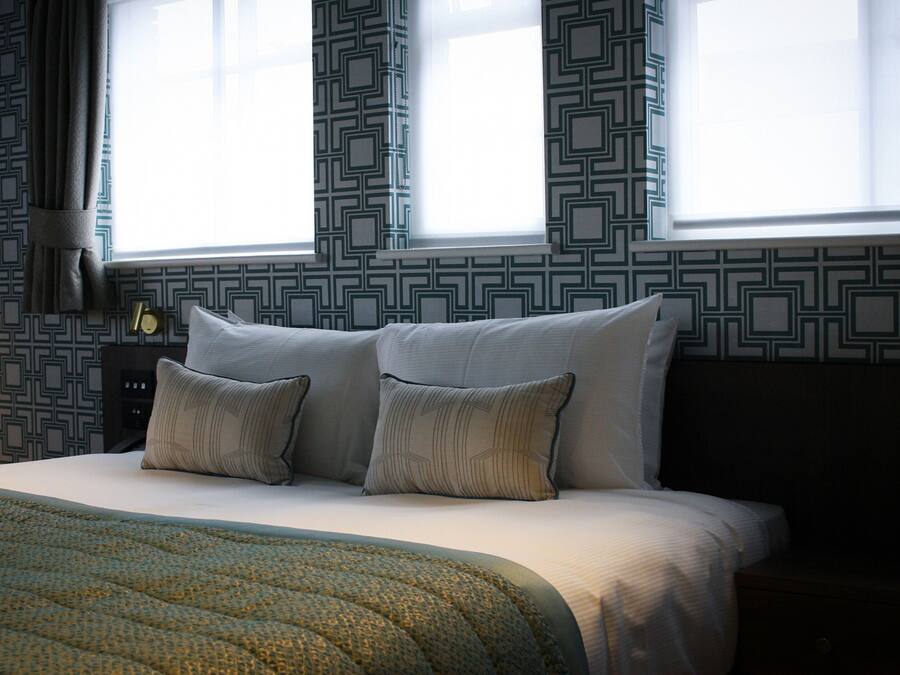 A neatly made bed with two pillows, a patterned wall, and a window with curtains.