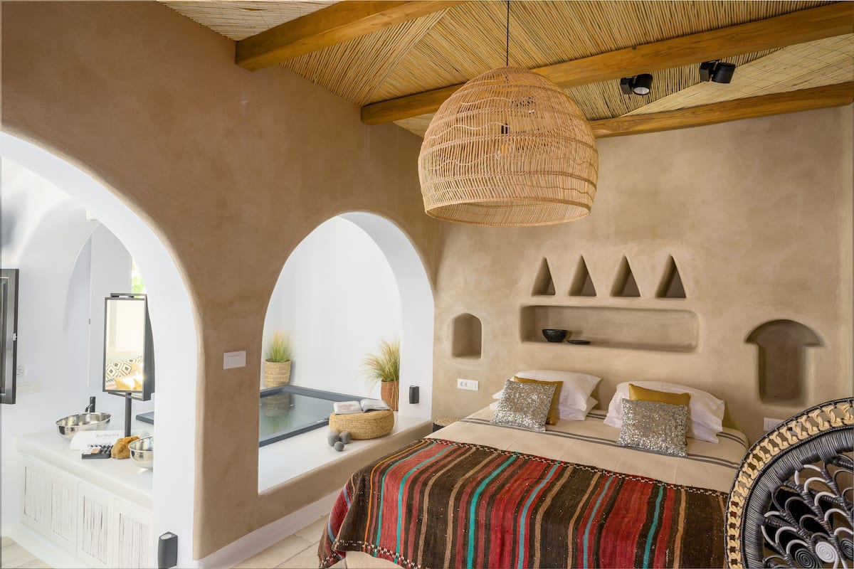 A bedroom with a bed, a woven ceiling light, and a view through an archway.