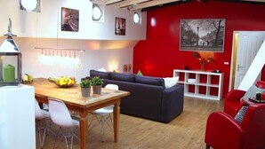 A modern living room with a wooden dining table, a dark blue sofa, and a red armchair. There are potted plants on the table and a white console table against the red wall. - Boathotel Rotterdam Seven (Rotterdam)