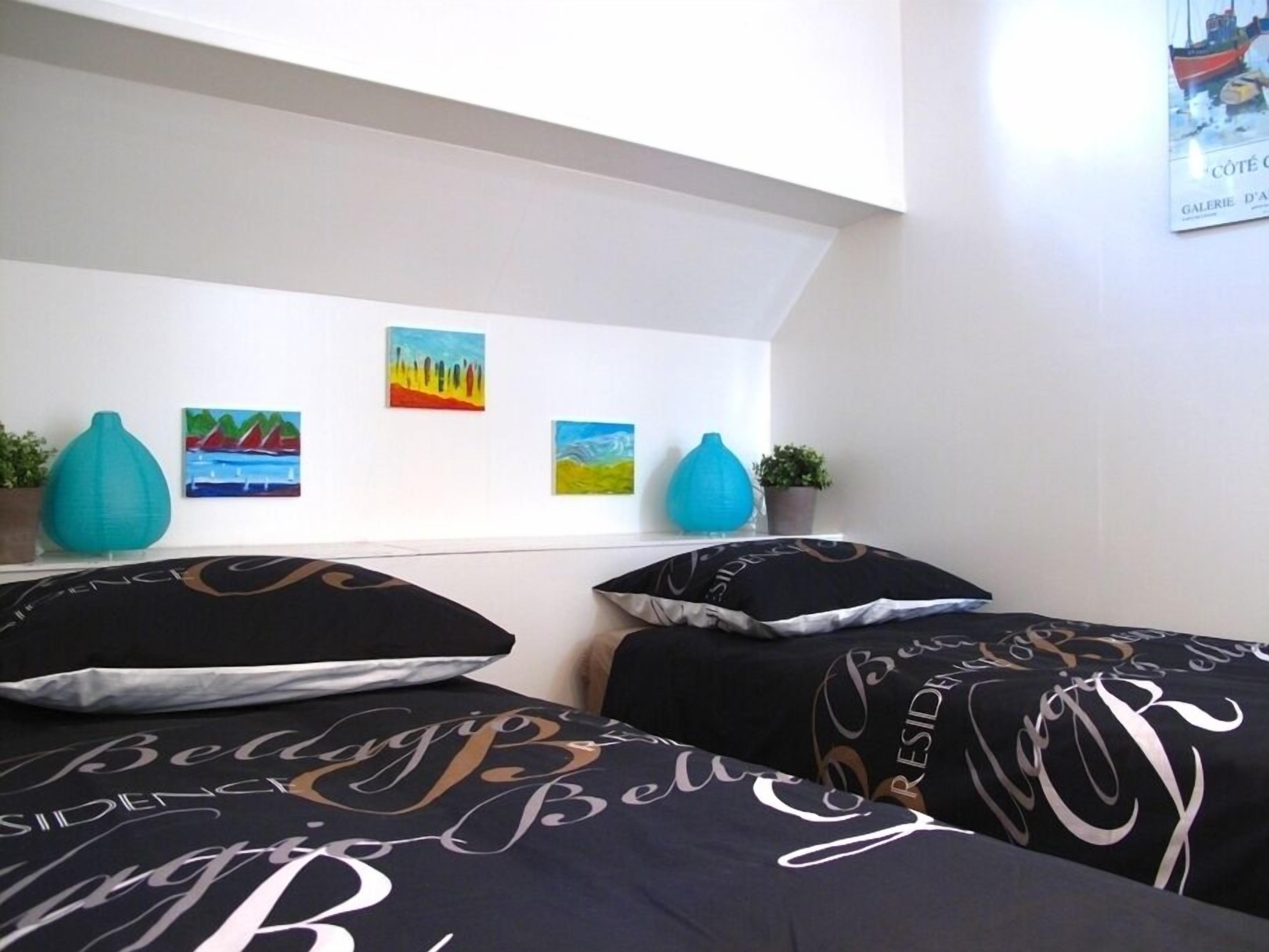 Une chambre avec deux lits, chacun agrémenté de draps à motifs noir et blanc. Trois tableaux sont accrochés au mur au-dessus des lits, et deux vases bleus sont posés sur la table de chevet.