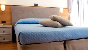 A bed with a blue quilt and two pillows, a wooden headboard, a bedside table with a lamp, and a window with curtains. - Hotel Marinella (Celle Ligure)