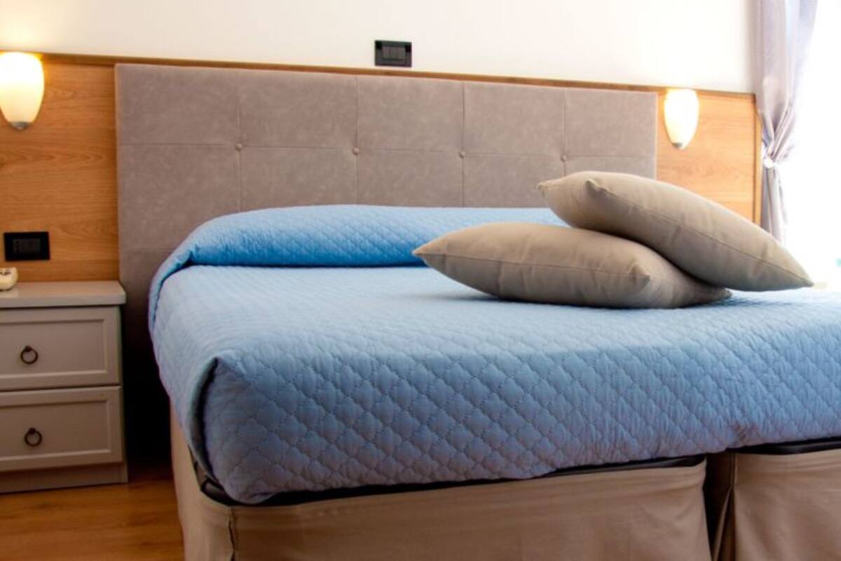 A bed with a blue quilt and two pillows, a wooden headboard, a bedside table with a lamp, and a window with curtains.