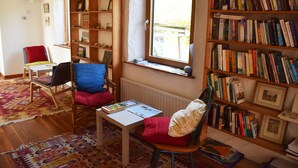A cozy reading nook with a wooden bookshelf, a comfortable chair, a small table, and a large window. - The Song House (Falcarragh)